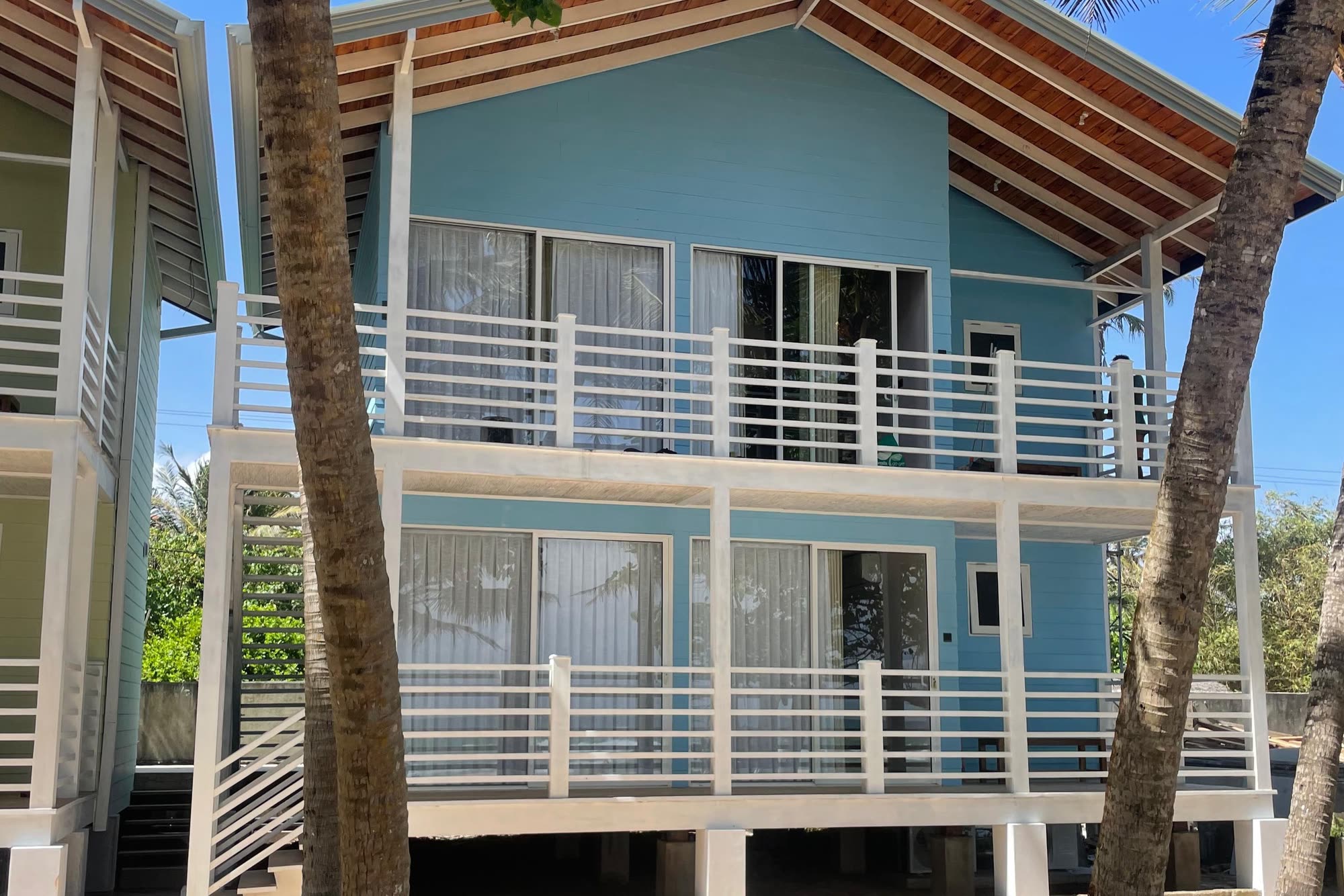 A pale blue two-storey timber chalet with white balconies and a timber-lined roof, framed by palm trunks under a blue sky