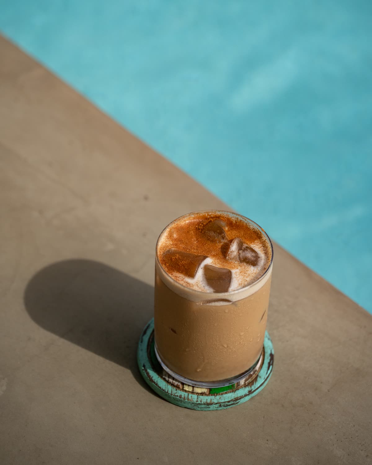 An iced drink dusted with spice on a turquoise coaster at the edge of the pool