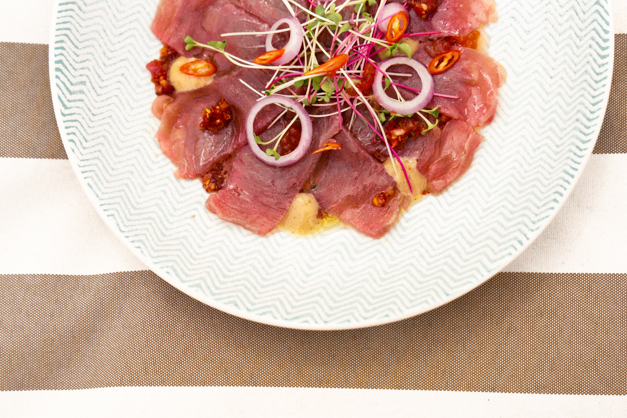 Thin slices of raw fish with red onion rings, sliced chilli and micro herbs on a white plate, on a brown and white striped towel