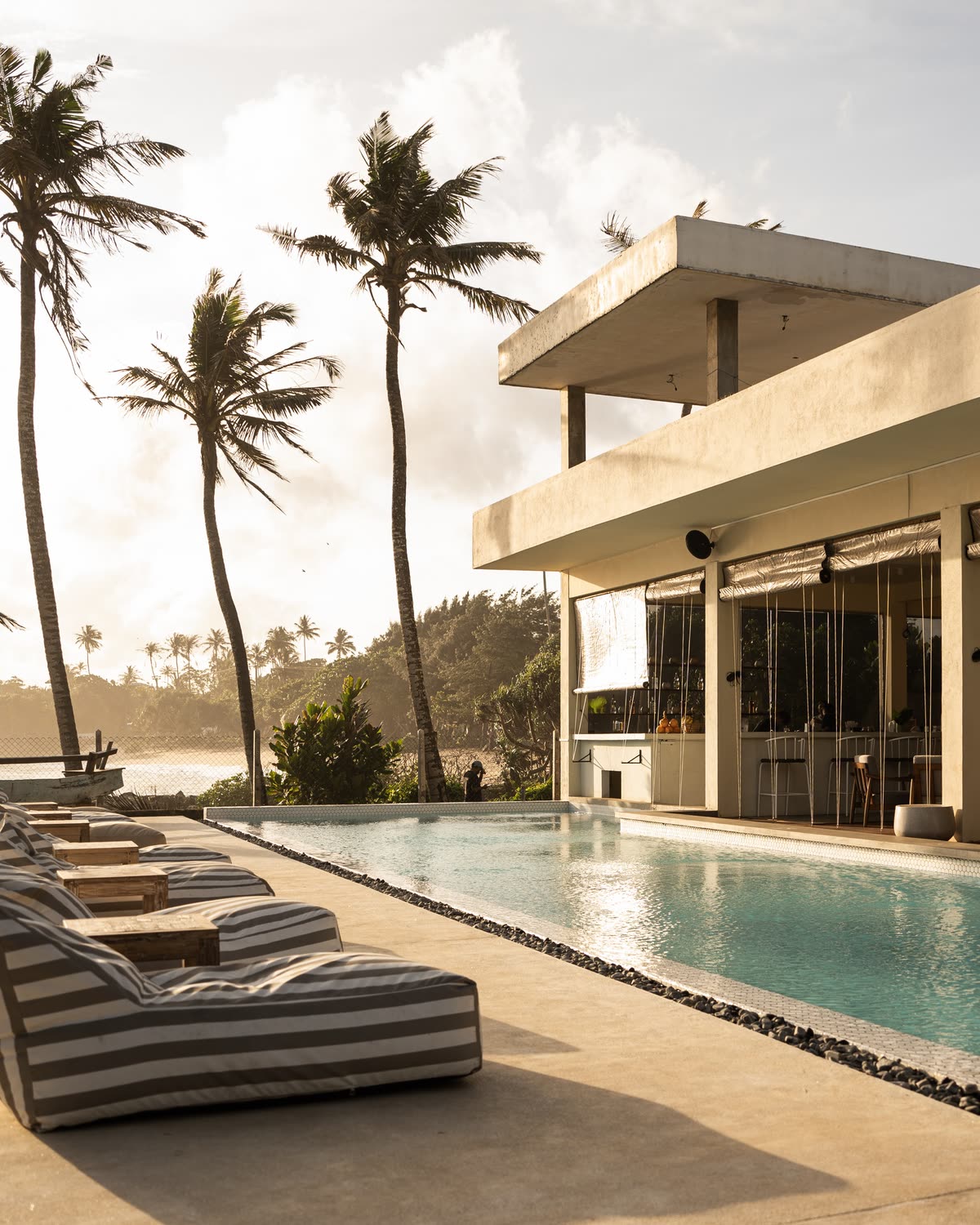 The white two-storey Club House beside the pool in late sun, tall palms and the coast to the left, striped bean bags along the deck