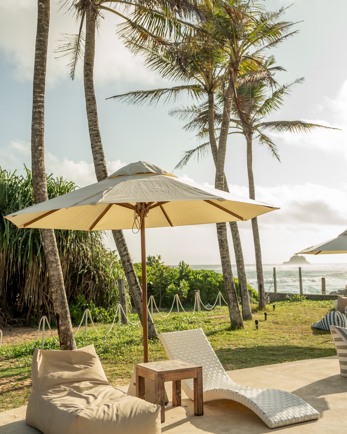 Tall palms over a cream parasol, a white woven lounger and a bean bag on the terrace, a small island on the sea beyond
